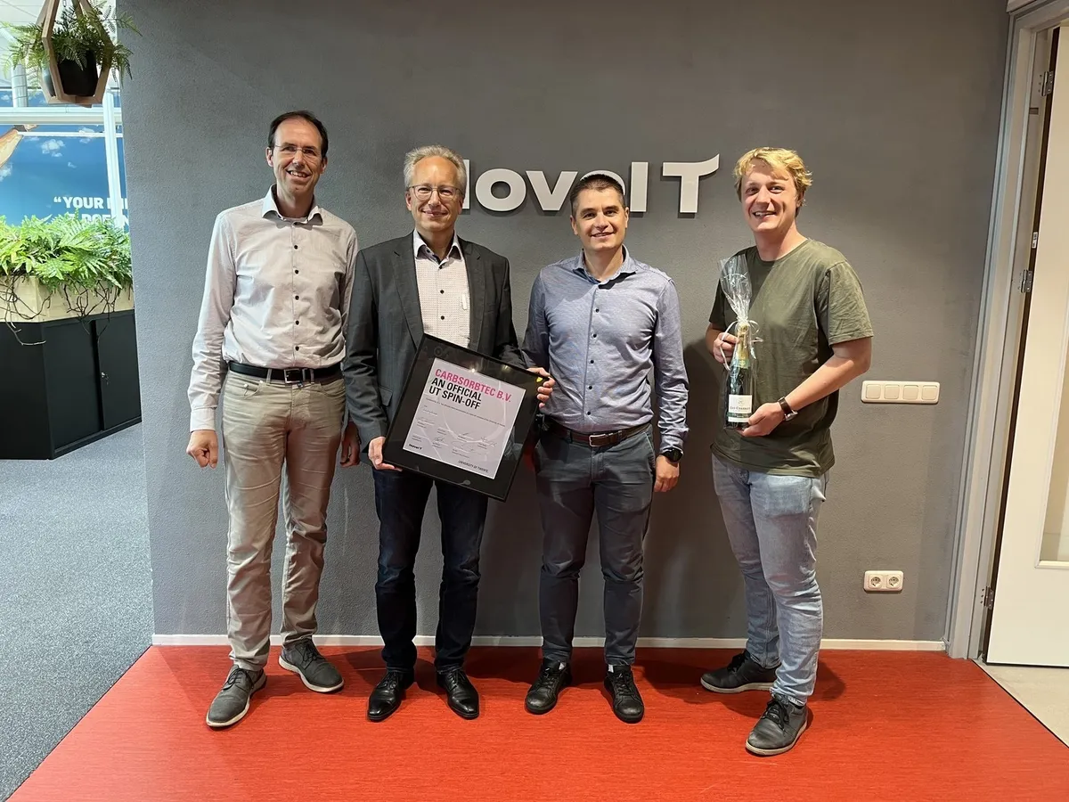 Four members of the CarbSorbTec team at Novel-T, holding a framed certificate naming CarbSorbTec B.V. an official University of Twente spin-off and a bottle of champagne